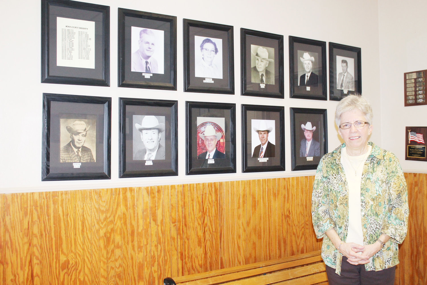 Teri Fox will retire at the end of January after working 38 years at the Wood County Sheriff’s Department. She worked for all  of the sheriffs whose photos are on the bottom row on the wall plus current Sheriff Jim Brown. (Monitor photo by Doris Newman)