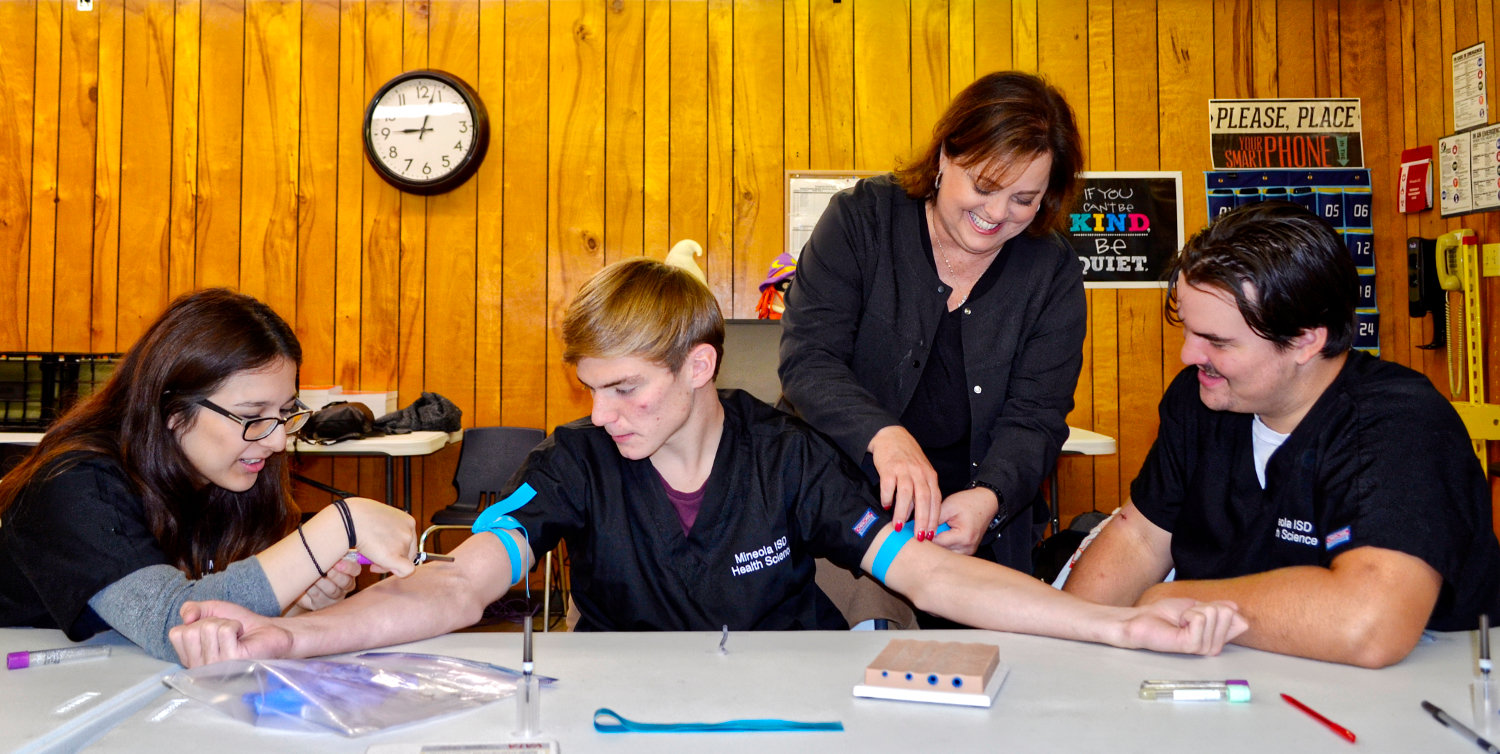 Students Anahi Aleman, Camden Adams and Dawson Elmore, in their third year of the health science program, get a lesson from instructor Cindy Robinson, RN, on how to feel for veins and correct needle positioning.